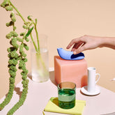 A minimalist composition highlights a hand reaching for a small, modern blue object—likely the sophisticated Dame Aer - Air Pulse Clit Stimulator by Dame—resting on a peach-colored cube. Nearby, there is a green glass, a white ceramic cup and saucer, a green plant in water, and a stack of yellow books set against a beige background.